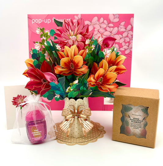 A 3D bouquet gift set with a variety of colorful artificial flowers, a pink candle, and a hand sanitizer bottle, all arranged in front of a pink envelope with floral designs.